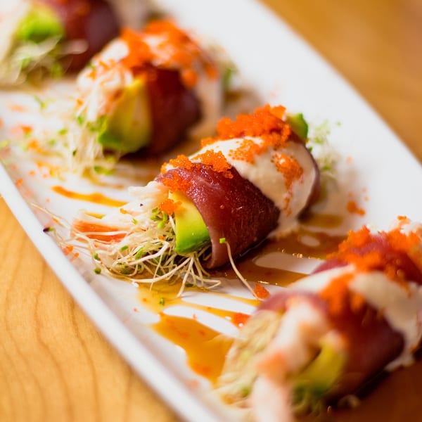 Four tuna and avocado rolls topped with white sauce, orange roe, and sprouts on a white plate.