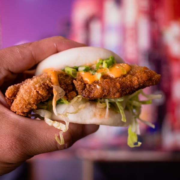 Hand holding a GoBistro fried chicken bao bun with lettuce and orange sauce.