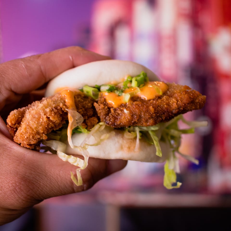 Hand holding a GoBistro fried chicken bao bun with lettuce and orange sauce.