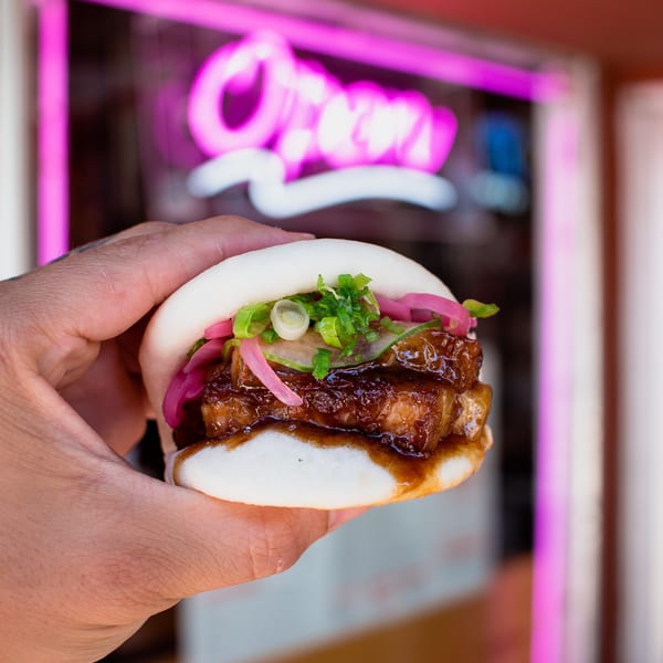 Hand holding a GoBistro pork belly bao bun with pickled onions and scallions, neon sign in background.
