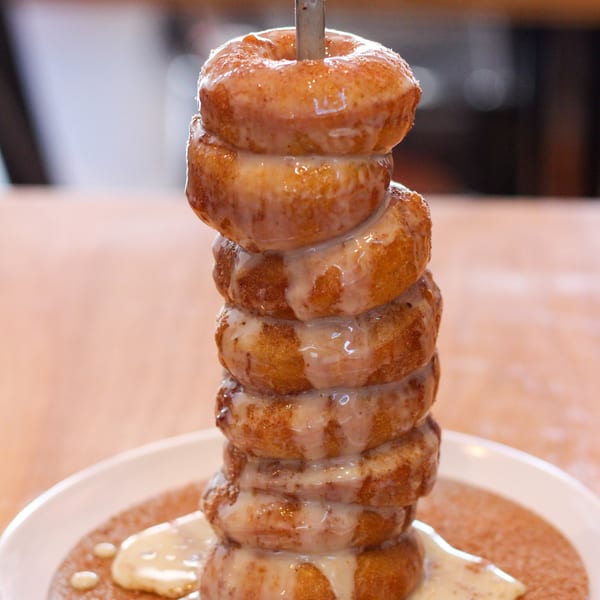 A tall stack of glazed donuts on a skewer, drizzled with icing and cinnamon sugar.